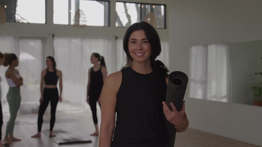 Woman smiles holding a rolledup yoga mat while others talk in the background