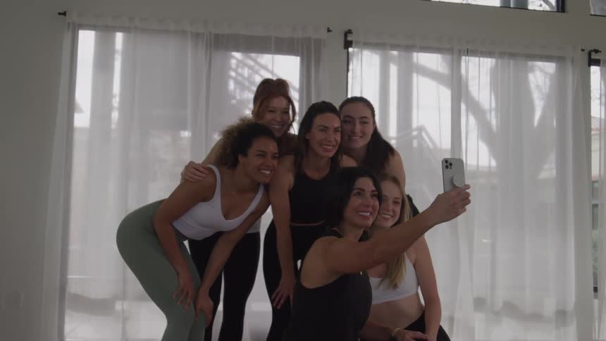 A diverse group of six smiling women in activewear takes a joyful selfie together in a brightly lit studio