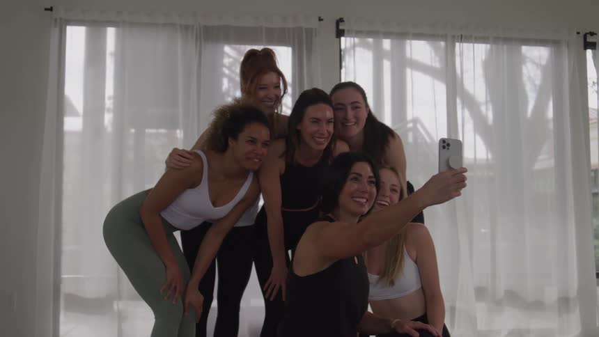 A group of six diverse women in athletic wear are gathered closely together in a bright studio smiling and posing for a selfie with a smartphone they appear to be friends or classmates celebrating a successful yoga or fitness session