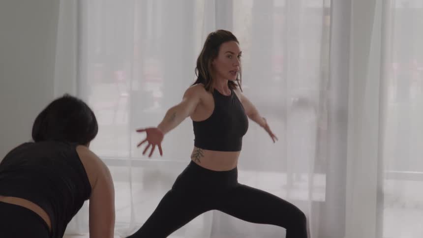 The video shows a woman performing a yoga pose likely a crescent lunge or a variation of it in a bright studio with other peopleshe starts with her hands near the floor in a low lunge then raises her torso and extends her arms overhead