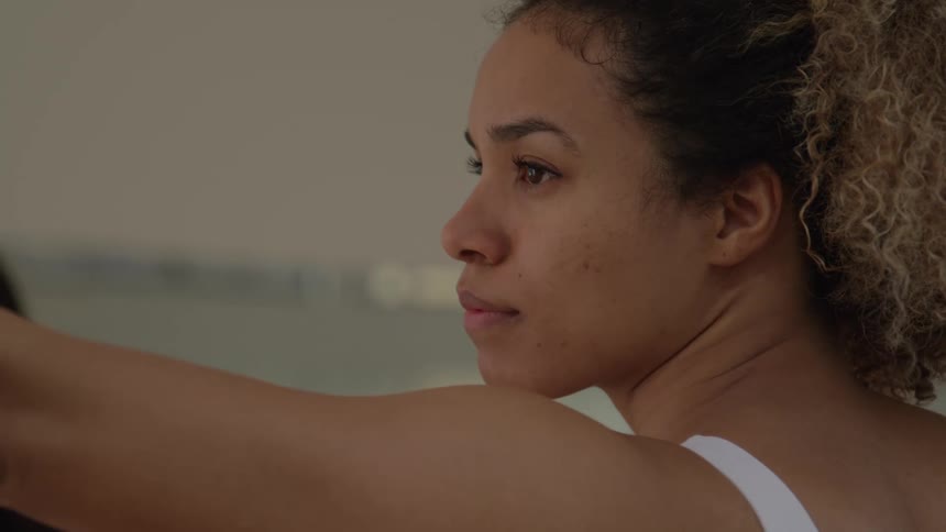 This is a closeup shot of a woman with curly hair who appears to be focused and engaged in an activity like yoga or stretchingshe is seen from the side with her arm extended forward and her expression suggests concentration and effort as she h