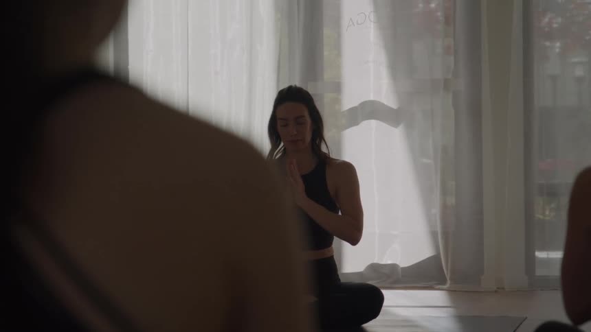 A woman in the foreground meditates in a cross legged position with her hands in prayer pose facing a bright window