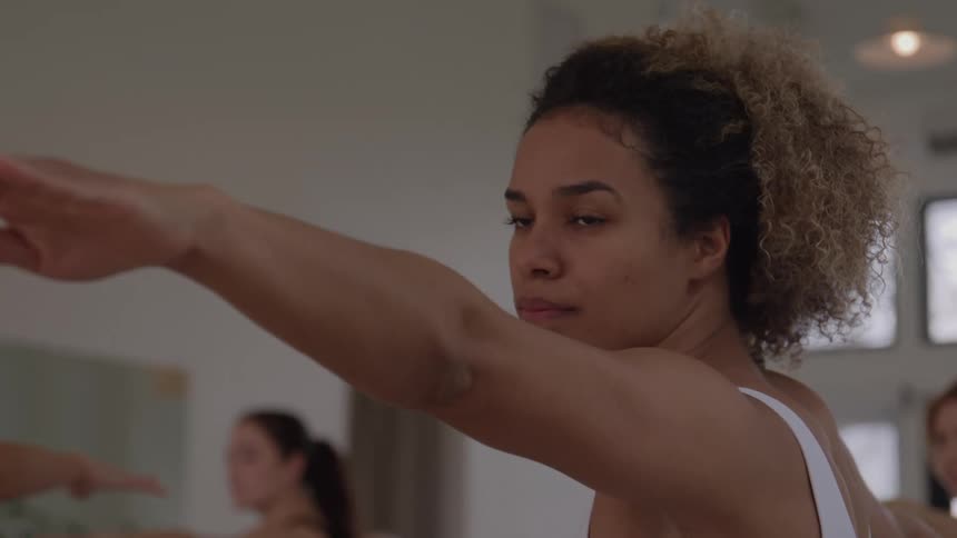 A focused woman with curly hair performs a yoga pose with outstretched arms looking intently forward in a bright studio