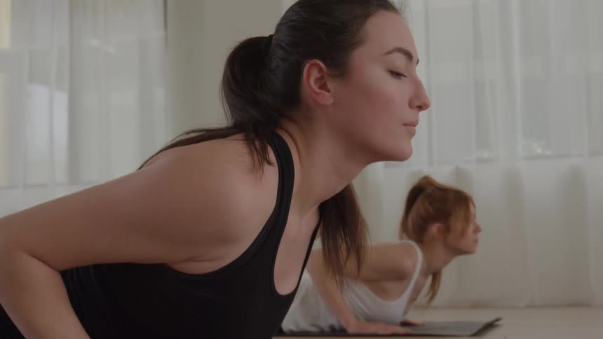 The video is a closeup shot of two women holding a yoga or fitness pose on their matsthey appear to be in the cobra pose bhujangasana or the upward phase of a yoga pushup lifting their chests off the floor with a focused expression