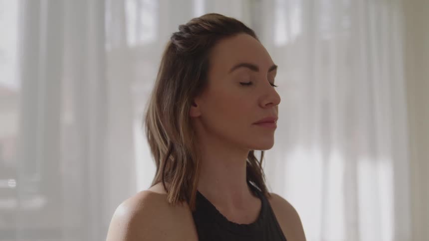 A serene woman with her eyes closed and a calm expression meditating peacefully in a brightly lit room