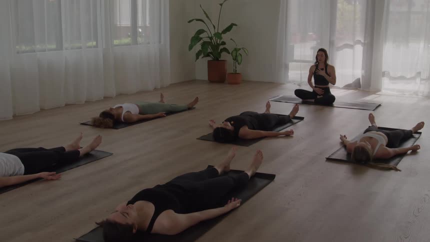 A female yoga instructor sits at the front of the class at midday while other women lie flat on their backs on yoga mats