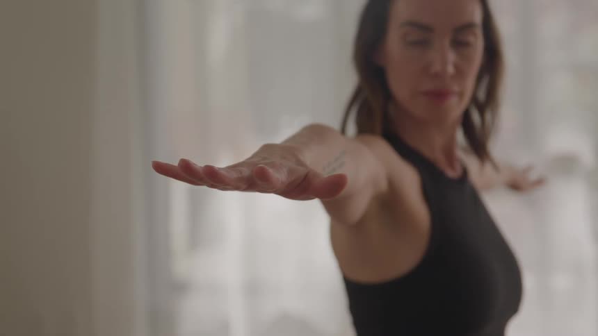 A young woman practices yoga stretches indoors at midday standing with both hands spread apart