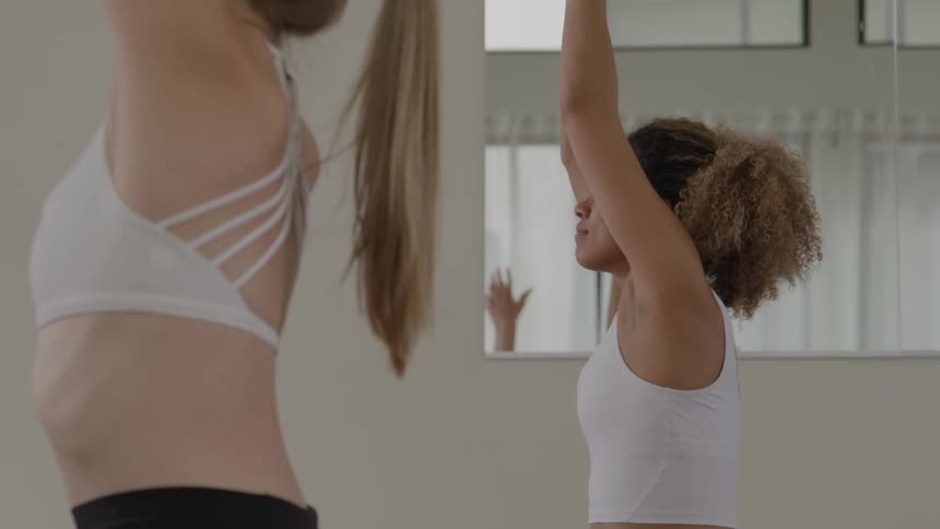 Two women standing sidebyside in a yoga studio beginning in a prayer pose then raising their arms overhead and finally lowering their hands back to the chest in the prayer pose