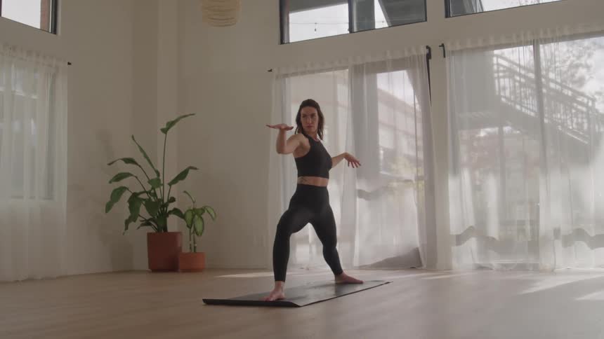 A young woman practices yoga stretches indoors at midday standing with both hands spread apart