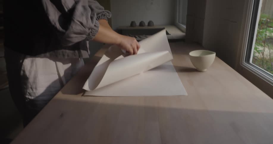 A young asian woman wrapping a small ceramic bowl with a sheet of paper on a table surface in a pottery room during daytime