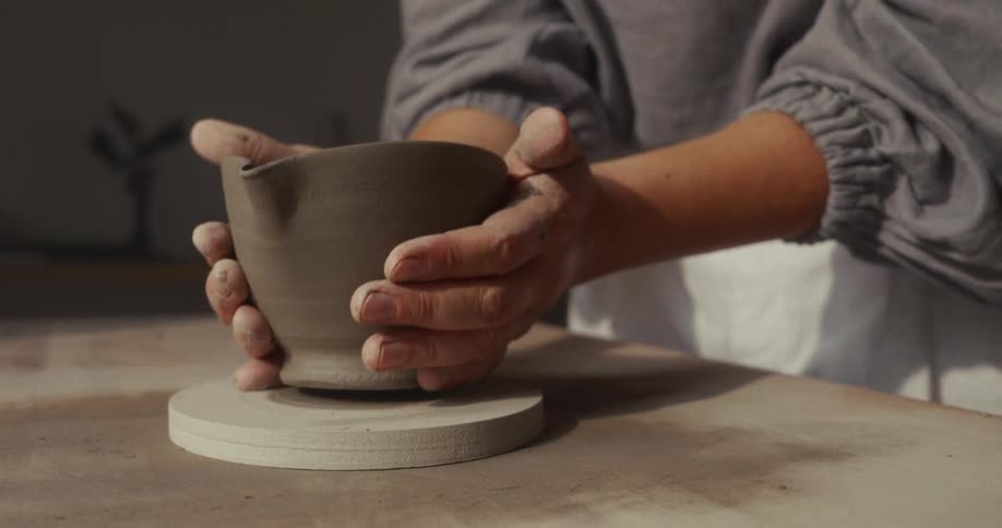 Close up of a young woman lifting a clay pottery cup from a tabletop in a room during daytime