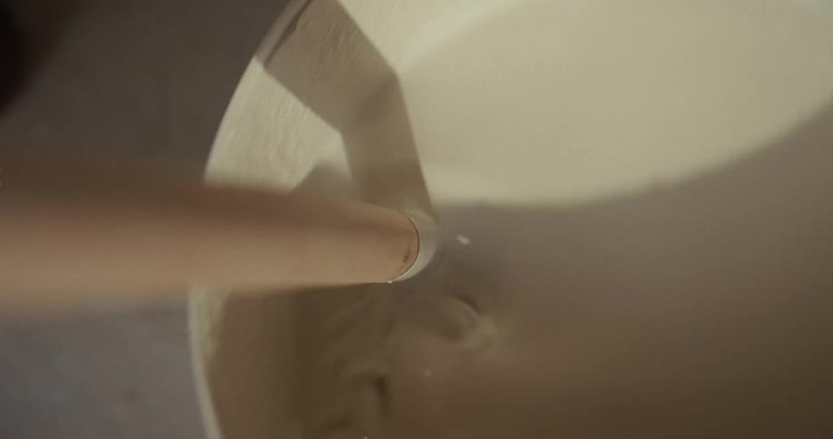 A person uses a long stick to stir white paint inside a white bucket at midday