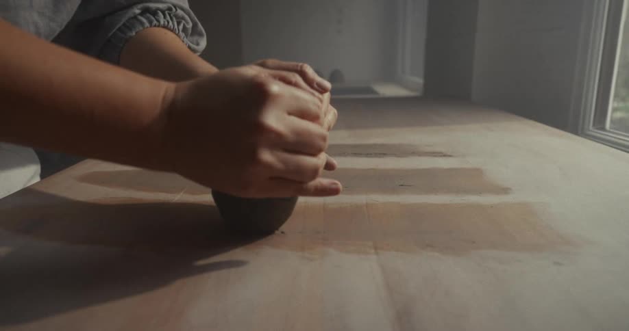 A persons hands pushing and rolling a lump of dark material on a wooden table near a window