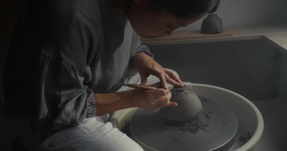 Close up of a young asian woman using a tool to finish a clay pottery on a pottery wheel in a room during daytime