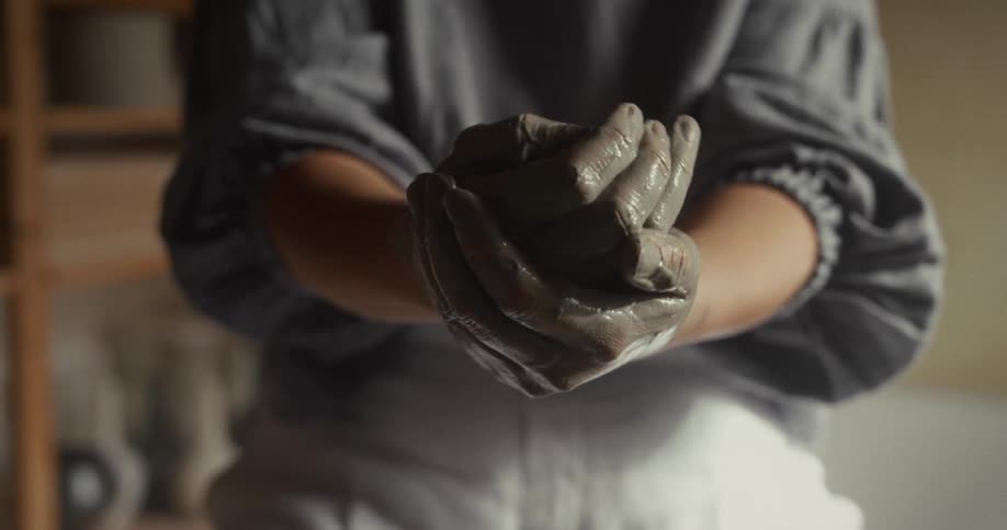 A potter rubs wet clay on her hands while sitting indoors