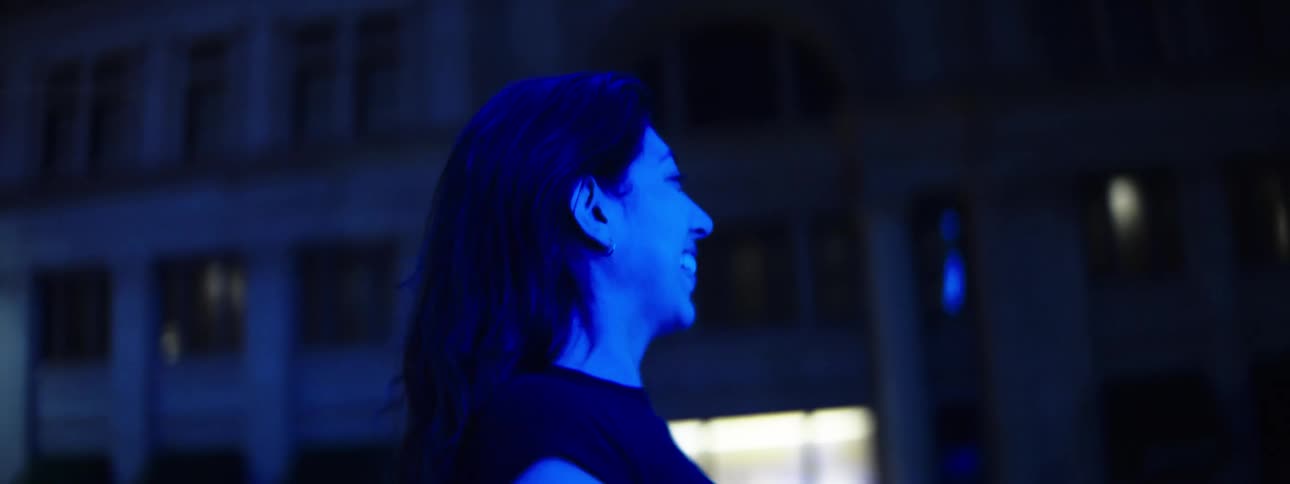 The profile of a woman with dark hair standing outdoors at night under blue light smiling while touching their hand to their face while a bus pass by her side