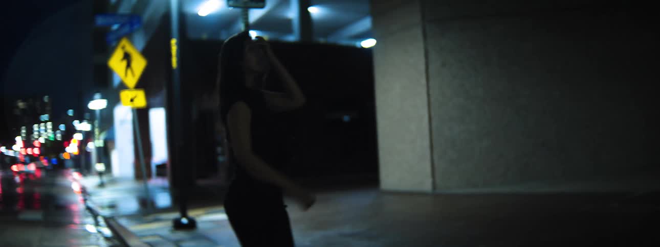A woman walks along a dark city street at night with streetlights and blurred car lights illuminating the scene