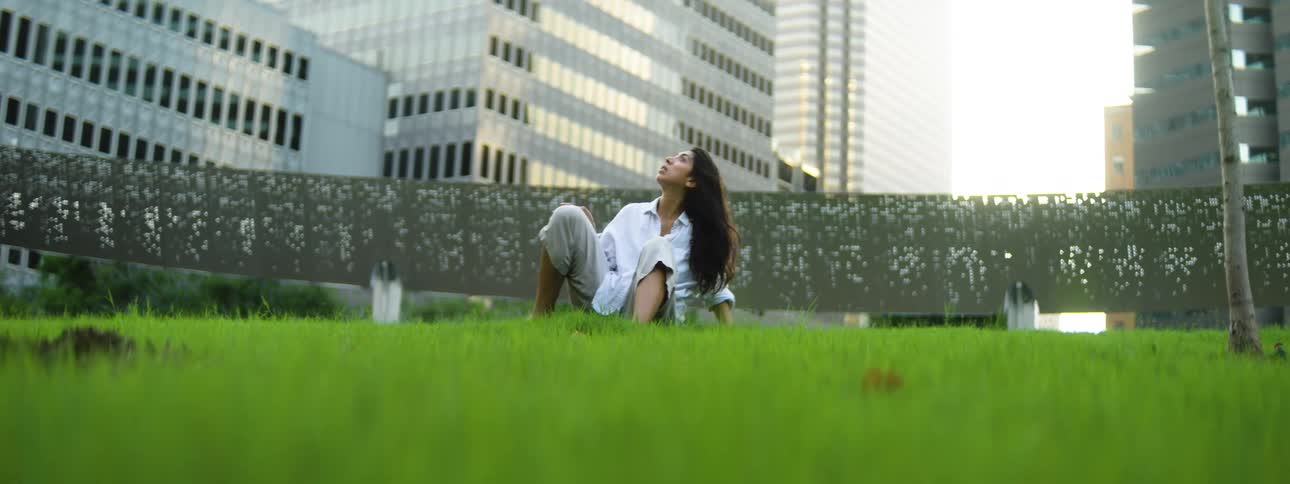 A hispanic woman with long dark hair sitting on a patch of grass in front of tall city buildings who then slowly moves their body from a kneeling position into a sweeping backbend with one arm raised toward the sky