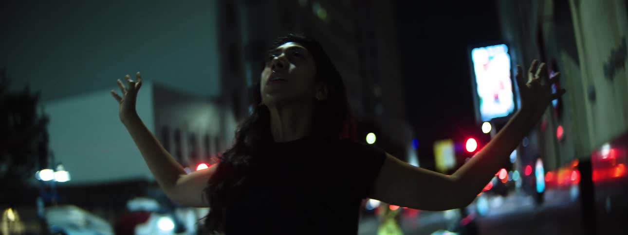 A hispanic woman with long dark hair wearing a dark top looks upward with a focused expression raising their arms out and walking on a city street at night
