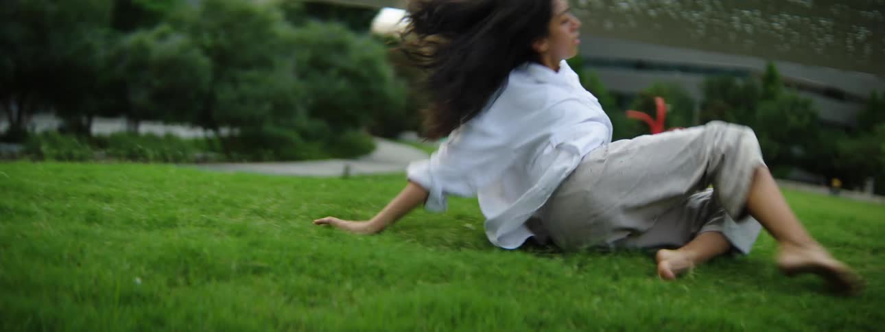 A female moving around erratically and dancing on a lawn surrounded by buildings