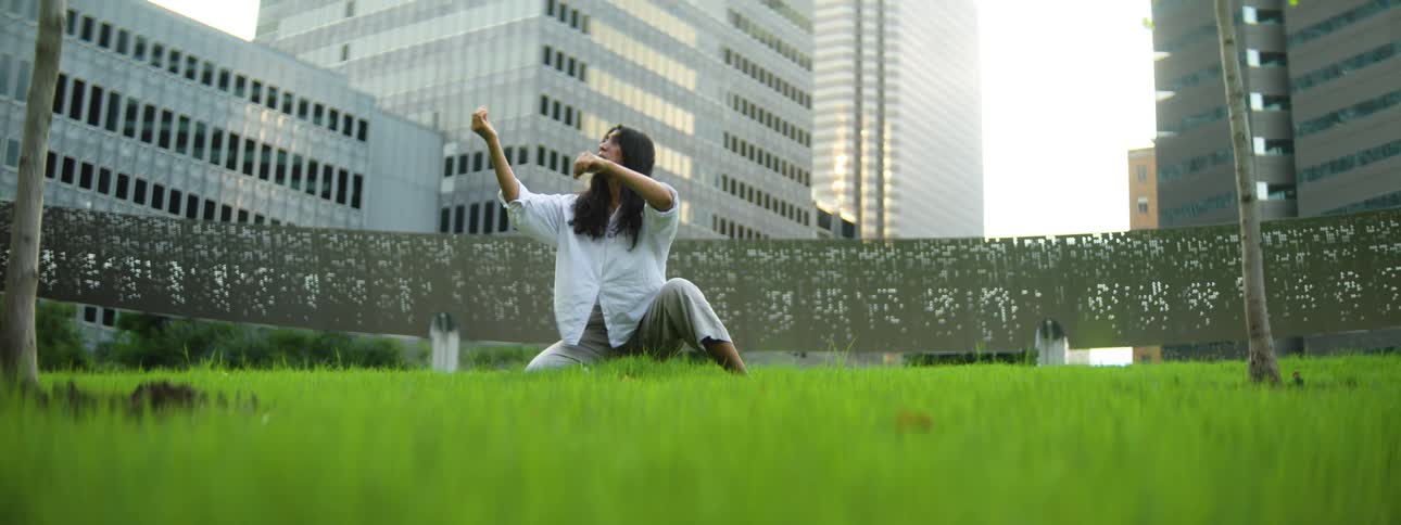 A woman in a loose white shirt practices a low focused stance on a bright lawn amid downtown towers