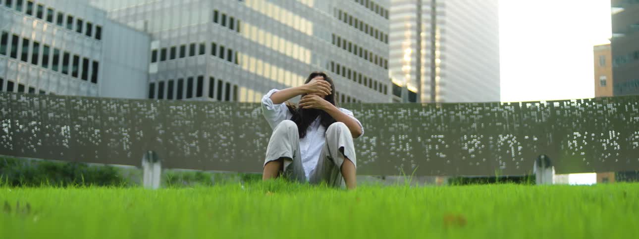 A hispanic woman with long dark hair wearing light colored clothes performs slow intentional movements from a prone position to a seated posture on a field of bright green grass with a backdrop of tall gridpatterned building