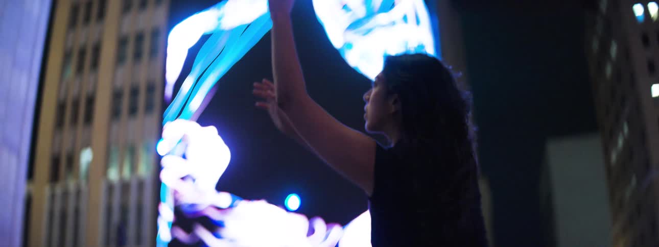 A woman at night gazes and dances near a towering abstract led display in a downtown plaza