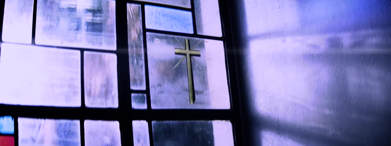A stained glass window with a golden cross set among geometric bluetoned panels glows with soft light