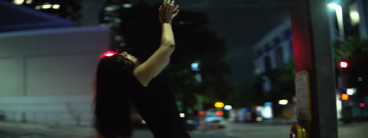 At a nightlit city intersection a woman in black leans back and raises her hands expressing urgent ecstatic energy