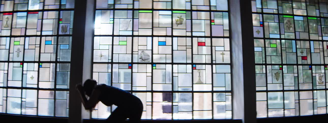 A woman with long hair dressed in dark attire stands in a large brightly lit framed window composed of numerous geometric and symbolic stainedglass panes
