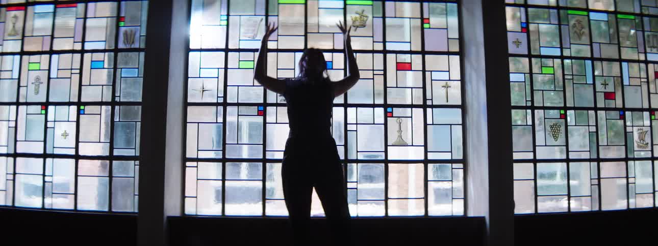 A hispanic young woman with long hair moves their body and arms in various poses and gestures against a large brightly lit stainedglass window