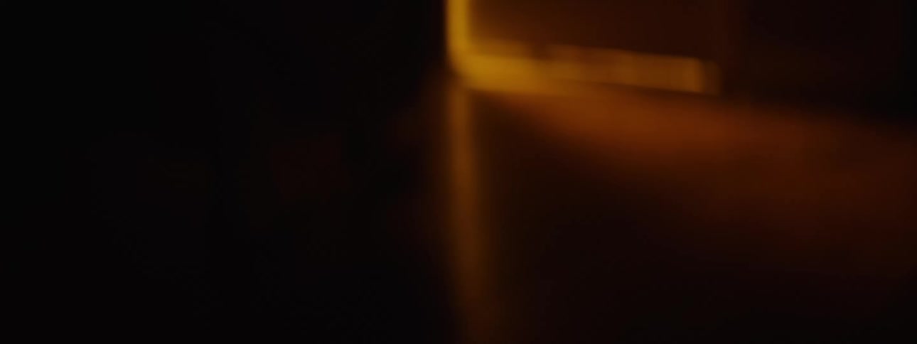 A person wearing wool socks and sleeping gown walking tentatively and slowly across a dark room towards a door with orange light coming from behind it then opens it slightly