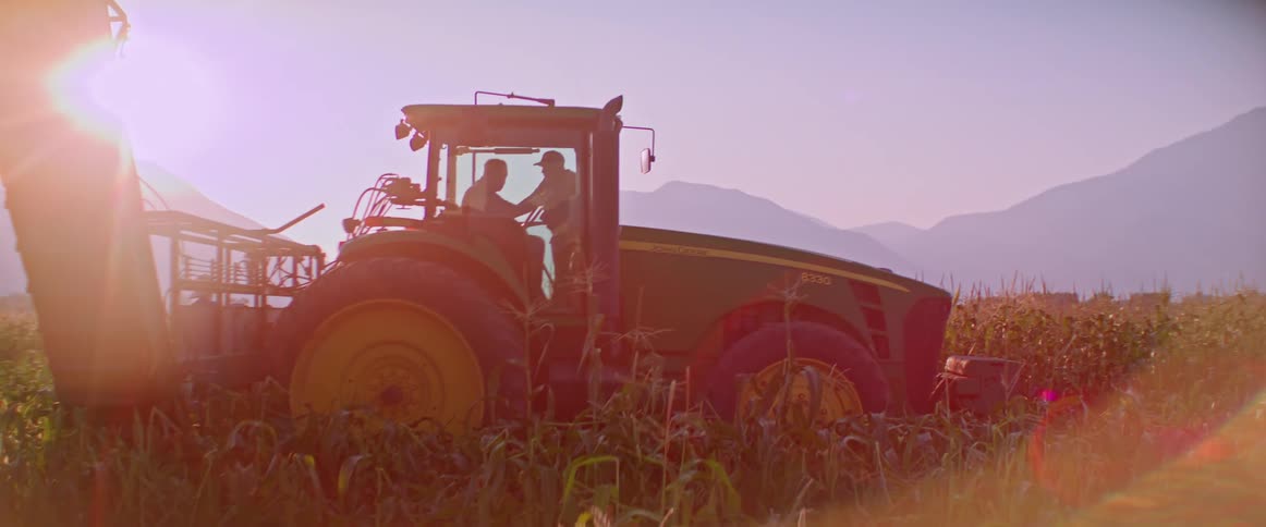 Two men operate a large green tractor with yellow wheels in a field of tall crops backdropped by mountains and a vibrant lens flare