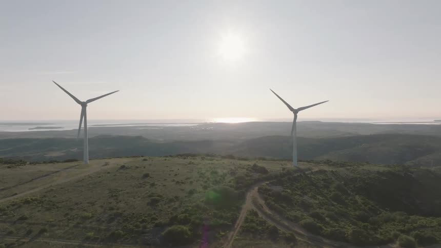 Two wind turbines majestically stand on a rolling hillside generating renewable energy under a bright hazy sky with distant water visible