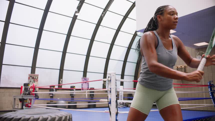 A focused female athlete with braided hair practices powerful tennis swings with a racket in a well lit gym with a boxing ring in the background