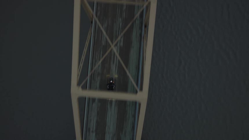 An overhead view shows a motorcycle man speeding across a long truss bridge with white supports over dark water or land at dusk or night