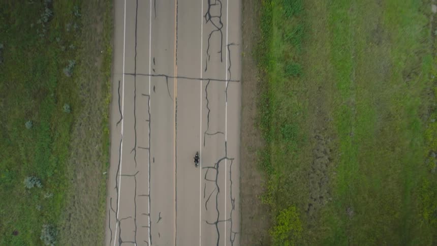 Overhead view shows a lone motorcyclist traveling a cracklined highway bordered by open grassy fields