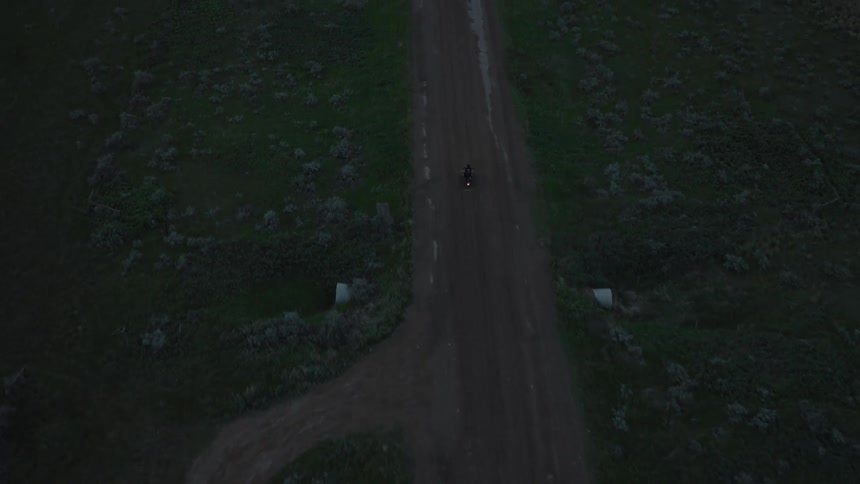 An aerial shot shows a lone figure on a bicycle riding along a dirt road that winds through a dark desolate landscape