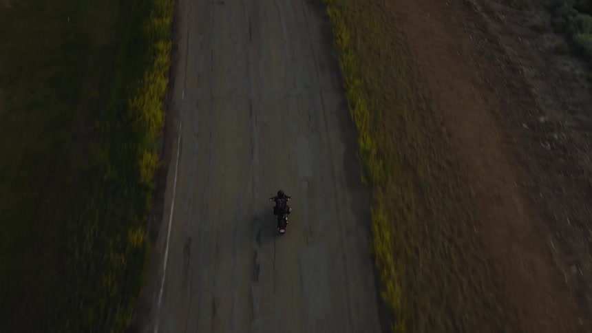 Aerial shot of a motorcyclist cruises a sunlit rural road casting a long shadow between grassy fields