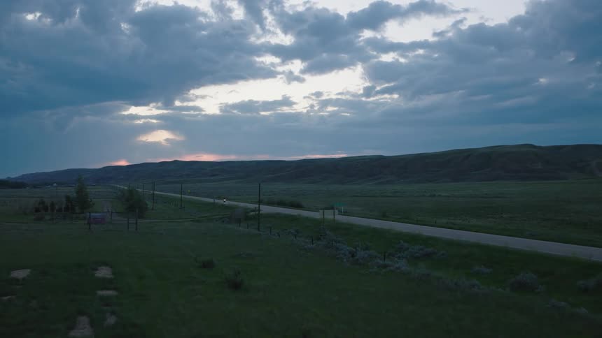 A motorcycle travels along a long rural road under brooding evening clouds as the last light glows on the horizon