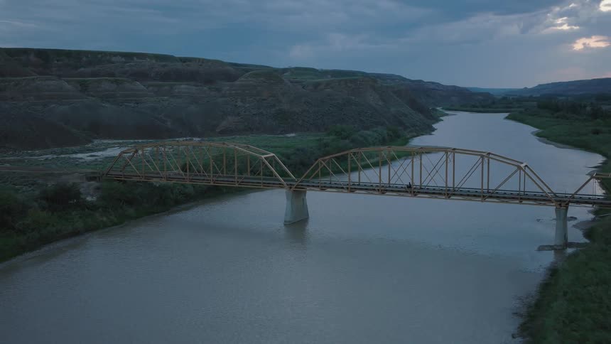 A motorcyclist is riding on an old truss bridge that spans a calm river at dusk with hills in the distance
