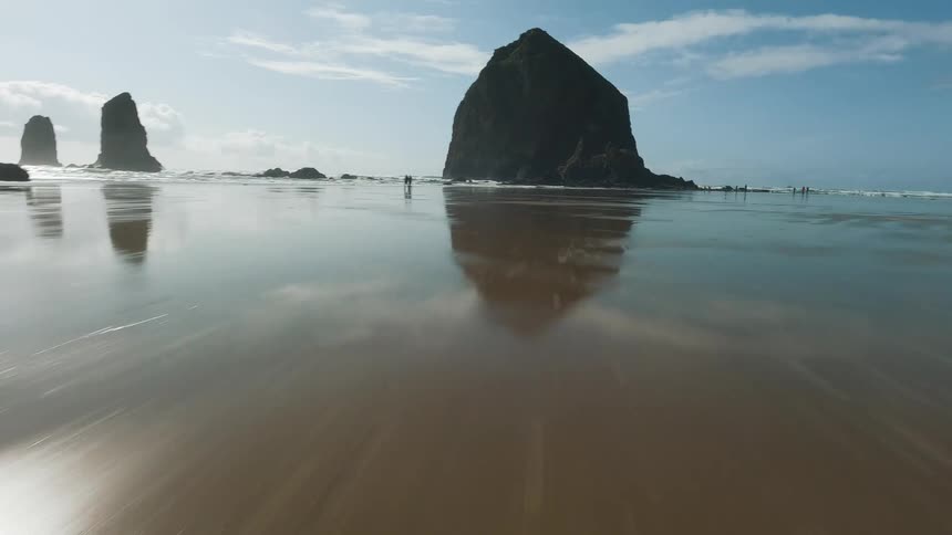 A wide view shows the wet reflective brown sand of a beach with haystack rock and other sea stacks rising from the ocean in the background with a person walking near the famous rock under a sunny sky
