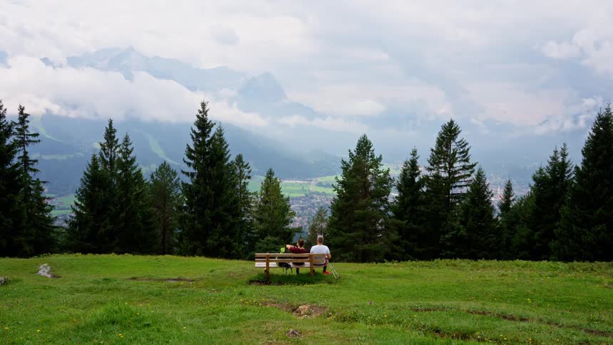 A man and a woman sit on a wooden bench on a high grassy hill enjoying a panoramic view of cloudcovered forested mountains and pine trees on an overcast day