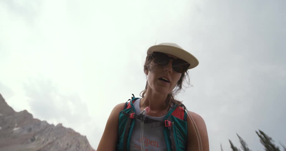 A young woman hikes along a mountain trail at midday wearing a hat a jacket and sunglasses under the bright sun