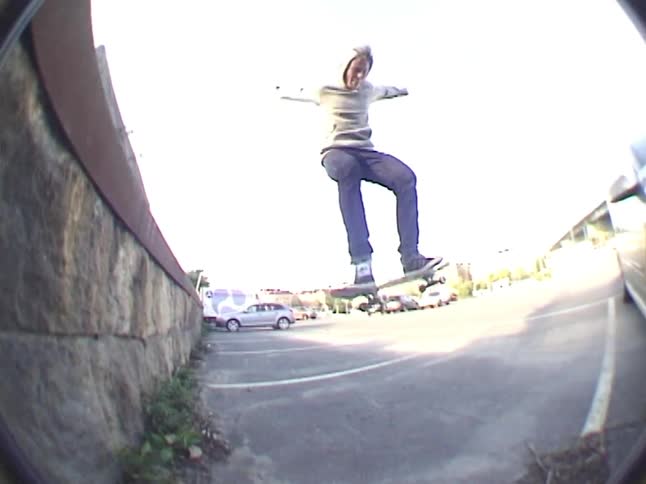 Hoodieclad skateboarder jumps over a large rock into a parking lot arms wide expression focused and exhilarated