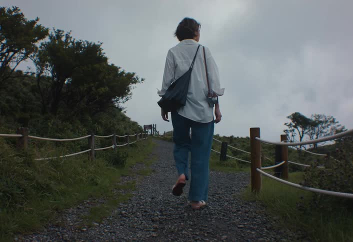 A young woman with short dark hair wears a white shirt and wideleg jeans with a shoulder bag while walking uphill on a gravel path bordered by a rope fence on a misty green hillside