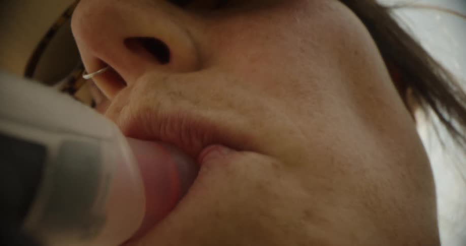 A young woman wearing glasses and a hat drinks water through a suction straw then spits it out