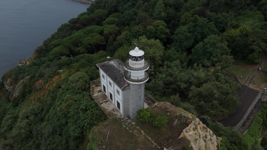 An aging cliffside lighthouse with attached keepers house overlooks dark calm water bordered by dense green trees