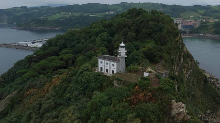 An old lighthouse with a house next to it on top of a mountain peninsula surrounded by forest trees