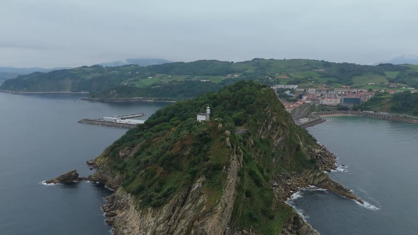 Cliff covered with vegetation and a lighthouse on top juts into the sea near a coastal town